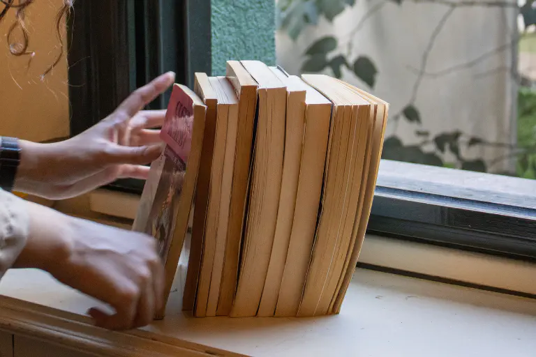 Mãos encostam uma pilha de livros a uma janela.