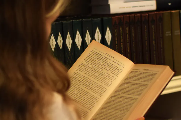 Uma mulher está em frente a uma estante e lê um livro.