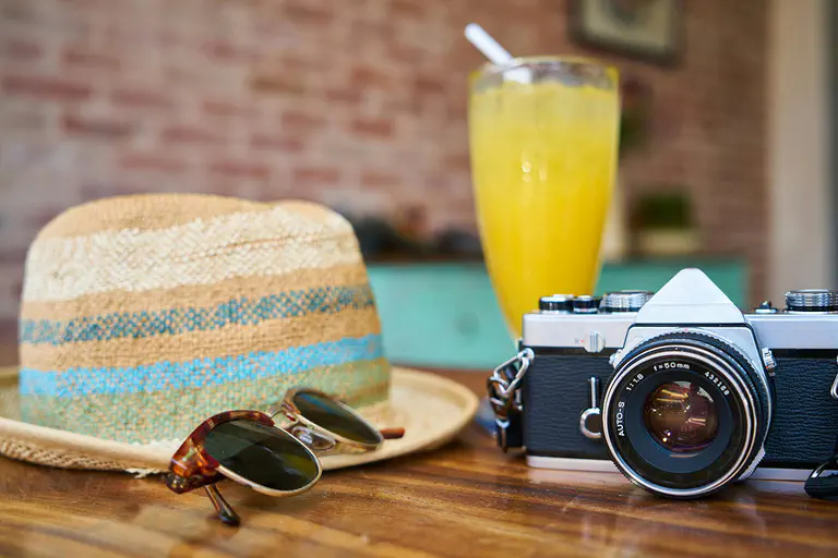 Vacation planning – picture shows sunglasses, an older camera, a straw hat and a glass of orange juice